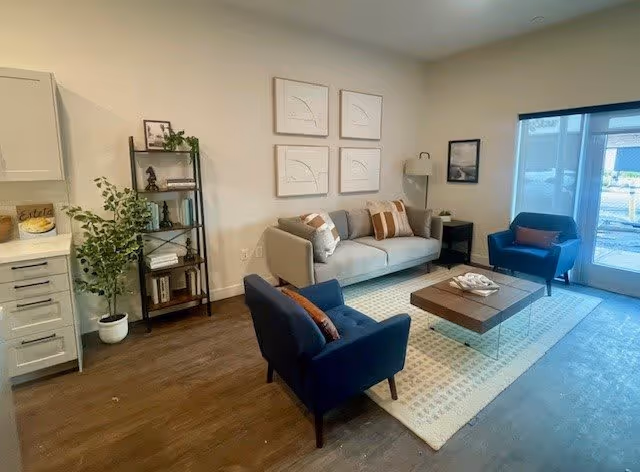 A cozy living room area with a gray sofa adorned with pillows, two blue armchairs with orange cushions, a wooden coffee table on a patterned rug, a black metal shelf with books and decorative items, a potted plant, and framed artwork on the walls. There is a large window or glass door letting in natural light.