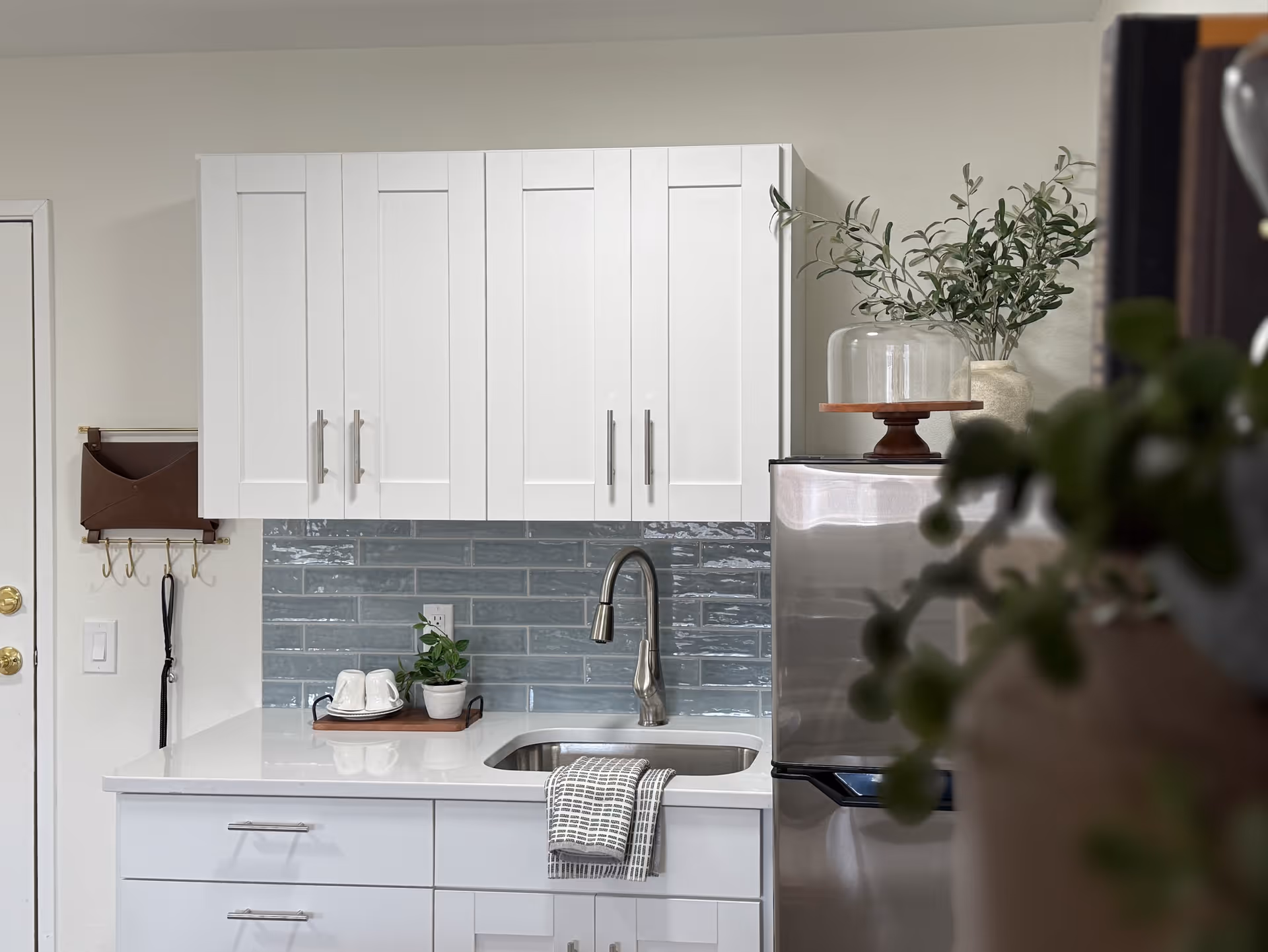 Small modern kitchen with white cabinets, stainless steel sink and refrigerator, blue tile backsplash, and countertop decor.