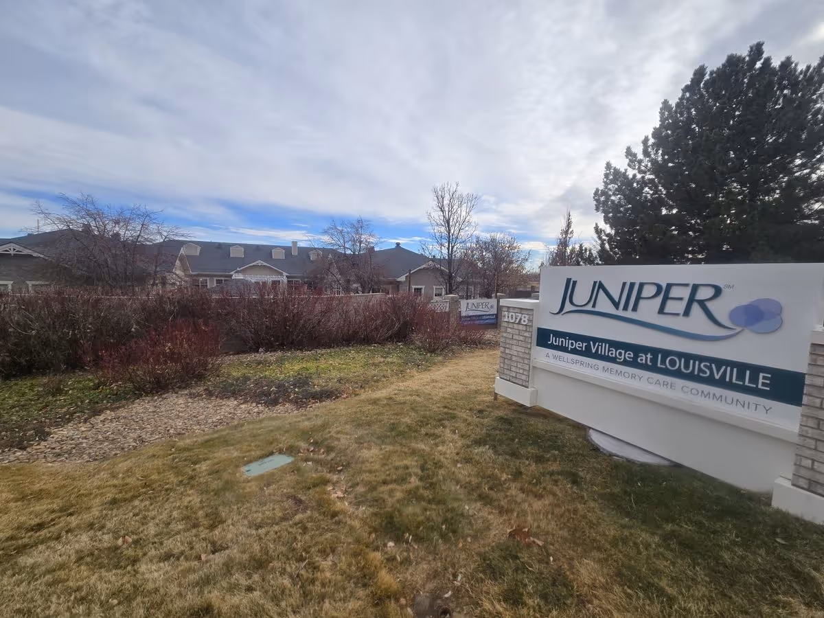 Entrance sign for Juniper Village at Louisville on a lawn with the facility buildings and trees in the background.