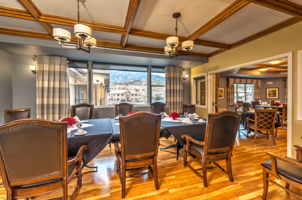 A dining room with wooden floors and a coffered ceiling featuring two chandeliers. The room has a large window with a view of buildings and mountains outside. There are multiple tables covered with black tablecloths, set with white napkins, cups, and small red floral centerpieces. The chairs are wooden with leather backs and seats. Another dining area is visible through an open doorway.
