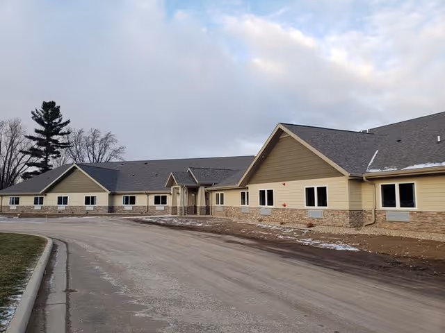 Exterior view of a single-story building with beige siding and stone accents, a sloped roof, and multiple windows. The building is situated alongside a paved road with some patches of snow on the ground and a cloudy sky overhead.