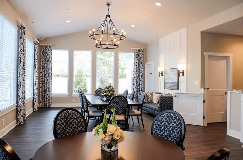 A bright and elegant dining room with large windows covered by patterned curtains, a round table with a floral centerpiece in the foreground, and a long rectangular dining table with chairs in the background. A modern chandelier hangs from the ceiling, and there is a seating area with a couch and wall sconces on the right side.