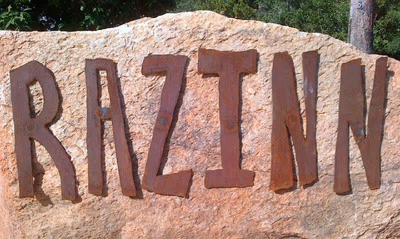 A large stone with the word 'RAZINN' spelled out in rustic, metal letters attached to its surface, set outdoors with greenery in the background.