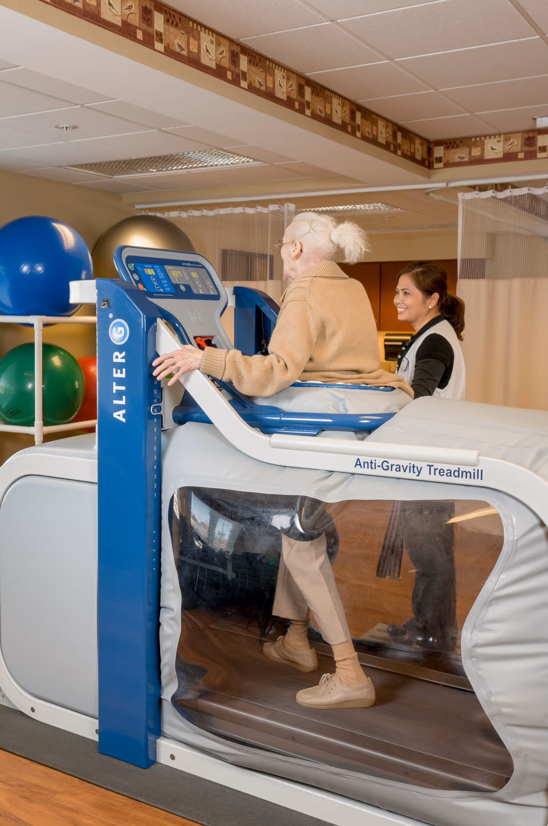 An elderly woman using an anti-gravity treadmill with the assistance of a healthcare professional in a rehabilitation or therapy room with exercise balls in the background.