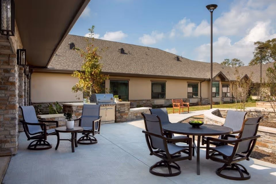 Outdoor patio area at The Shores at Clear Lake featuring multiple seating arrangements with cushioned chairs around round tables, a built-in stone grill, small plants on tables, and a well-maintained garden with trees and shrubs under a partly cloudy sky.