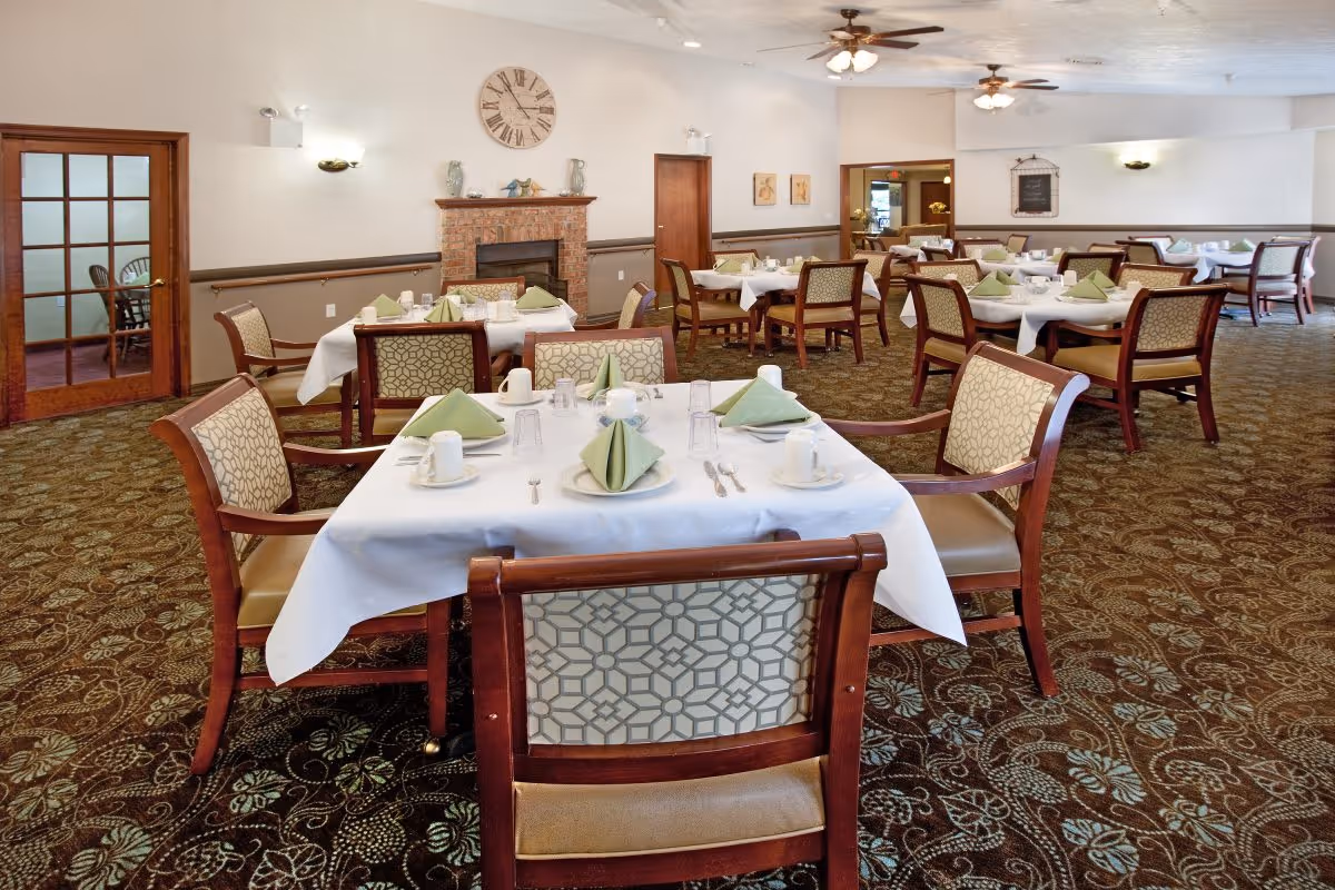 A spacious dining room with multiple tables covered in white tablecloths, each set with green folded napkins, cups, plates, and silverware. The room features patterned carpet, wooden chairs with cushioned seats and backs, a brick fireplace with a large clock above it, and ceiling fans with lights. There are wooden doors and framed artwork on the walls.