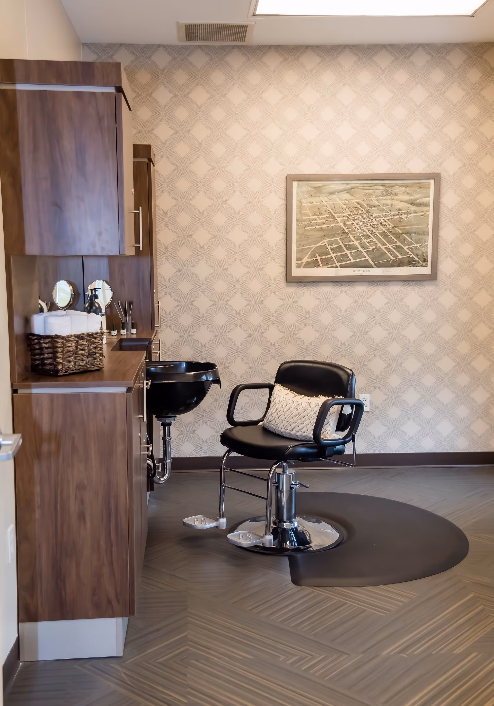 Interior view of a salon area in a nursing and rehabilitation center featuring a black salon chair with a decorative pillow, a black wash basin, wooden cabinetry with towels and grooming supplies, and a framed map on a patterned wallpapered wall.