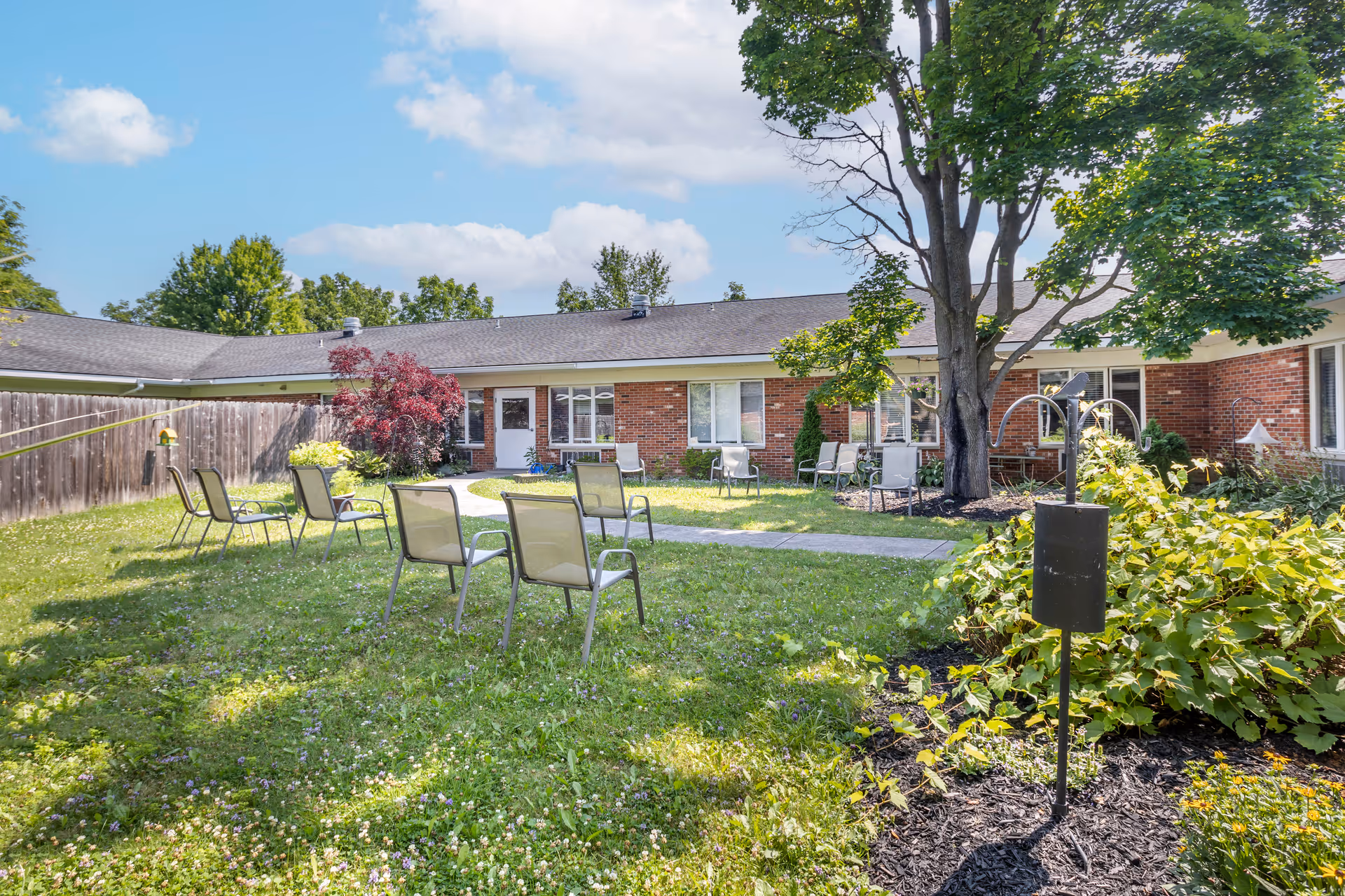 Outdoor courtyard area of a senior living facility with several metal chairs arranged on a grassy lawn. The courtyard is surrounded by a single-story brick building with windows and doors. There are trees, shrubs, and a wooden fence enclosing the space under a partly cloudy blue sky.