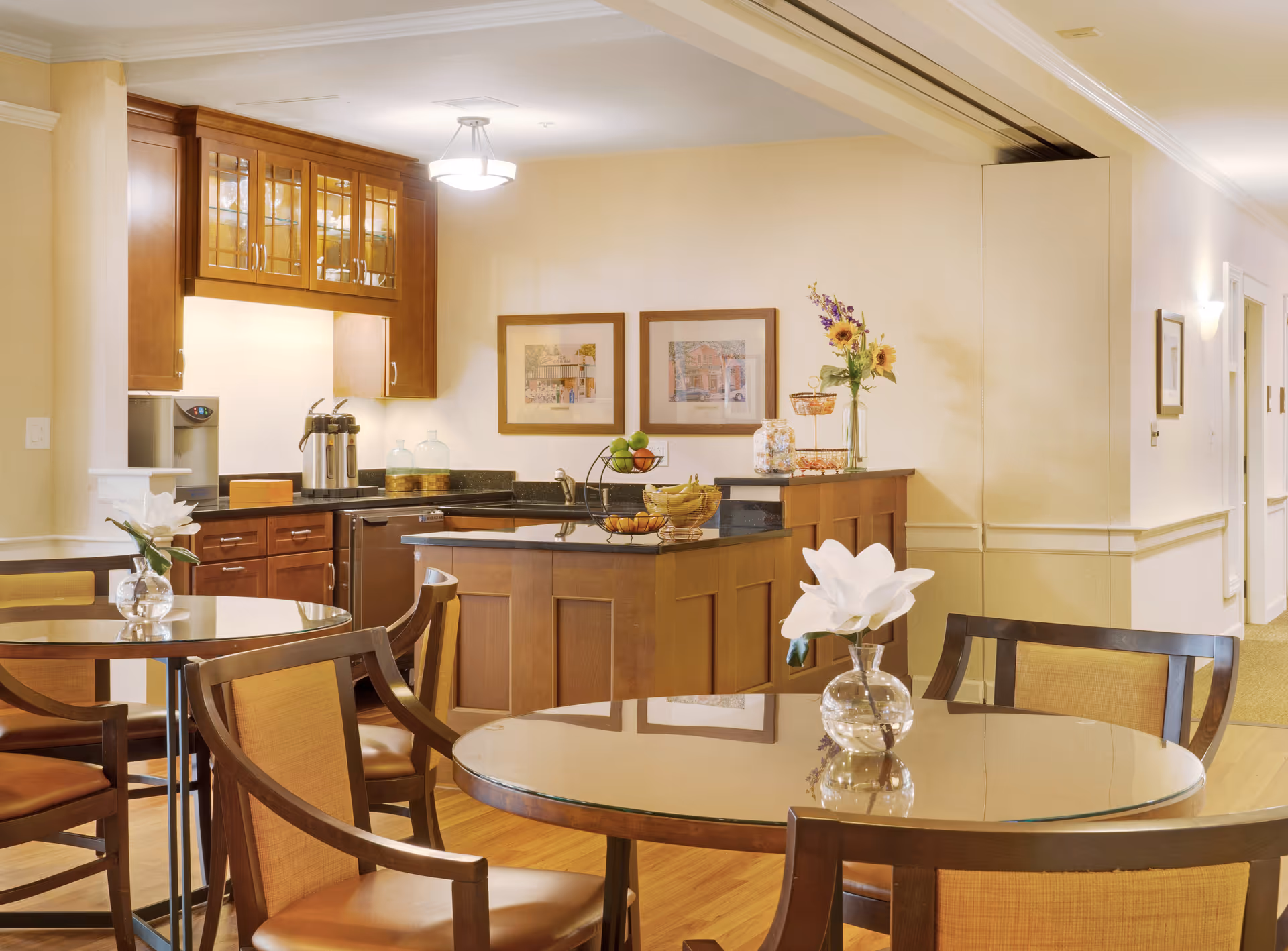 Bright communal dining area with round tables and chairs in front of a small serving kitchen and countertop decorated with fruit and flowers.