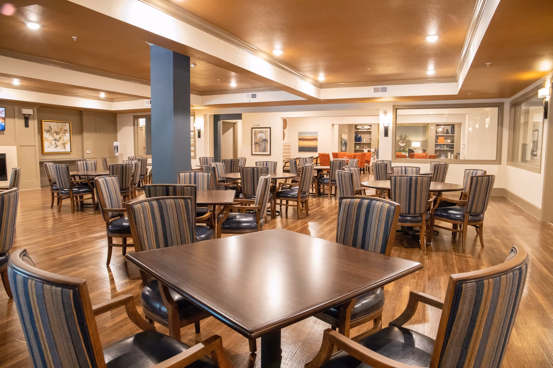 A spacious dining room with multiple wooden tables and striped cushioned chairs arranged neatly on a polished wooden floor. The room has warm lighting from ceiling fixtures and wall sconces, with framed artwork on the walls and a large window looking into another room with orange chairs and shelves.