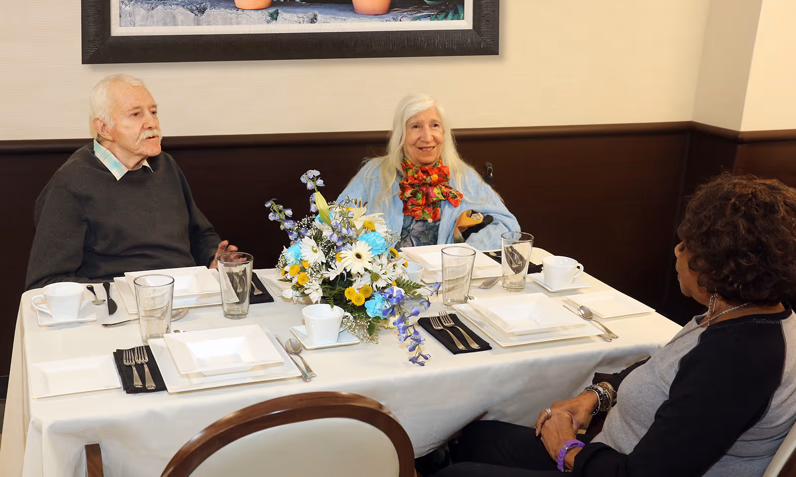 Three elderly people sitting around a dining table set with white plates, cups, glasses, and silverware. A floral centerpiece with blue, white, and yellow flowers is in the middle of the table. The room has beige walls with dark wood paneling and a framed picture hanging above.
