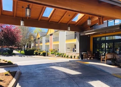 View of the exterior entrance of Jennings McCall Center with a covered driveway, benches, and a two-story building with balconies. Trees and shrubs line the walkway under a clear blue sky.
