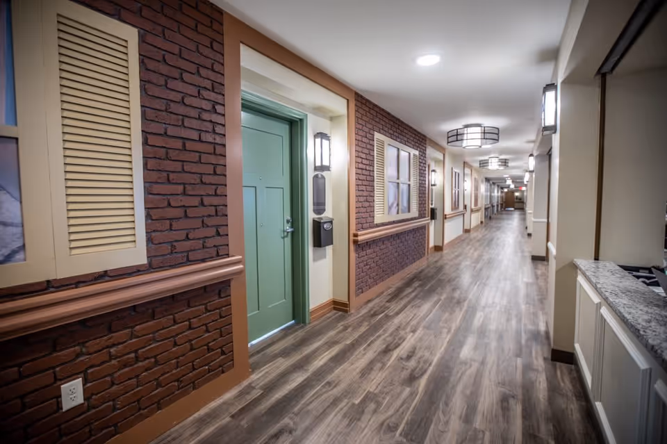 A long, well-lit hallway in a senior living facility with wood-patterned flooring, brick accent walls, green doors, and decorative wall sconces. The hallway features handrails and a reception counter on the right side.