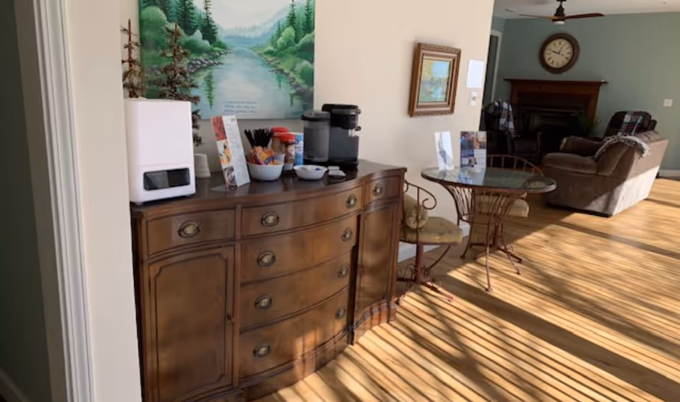 A cozy interior space with a wooden sideboard holding a coffee maker, cups, and snacks. Next to it is a small round glass table with two metal chairs. In the background, there is a living area with a sofa, armchairs, a fireplace, and a large clock on the wall. Sunlight streams through window blinds, casting shadows on the wooden floor.