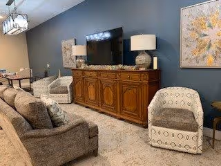 A cozy living room with a gray sofa and two patterned armchairs arranged around a wooden cabinet with a flat-screen TV on top. The walls are painted blue and decorated with framed artwork. Two table lamps with white shades sit on the cabinet, and a chandelier hangs from the ceiling. A carpet covers the floor.