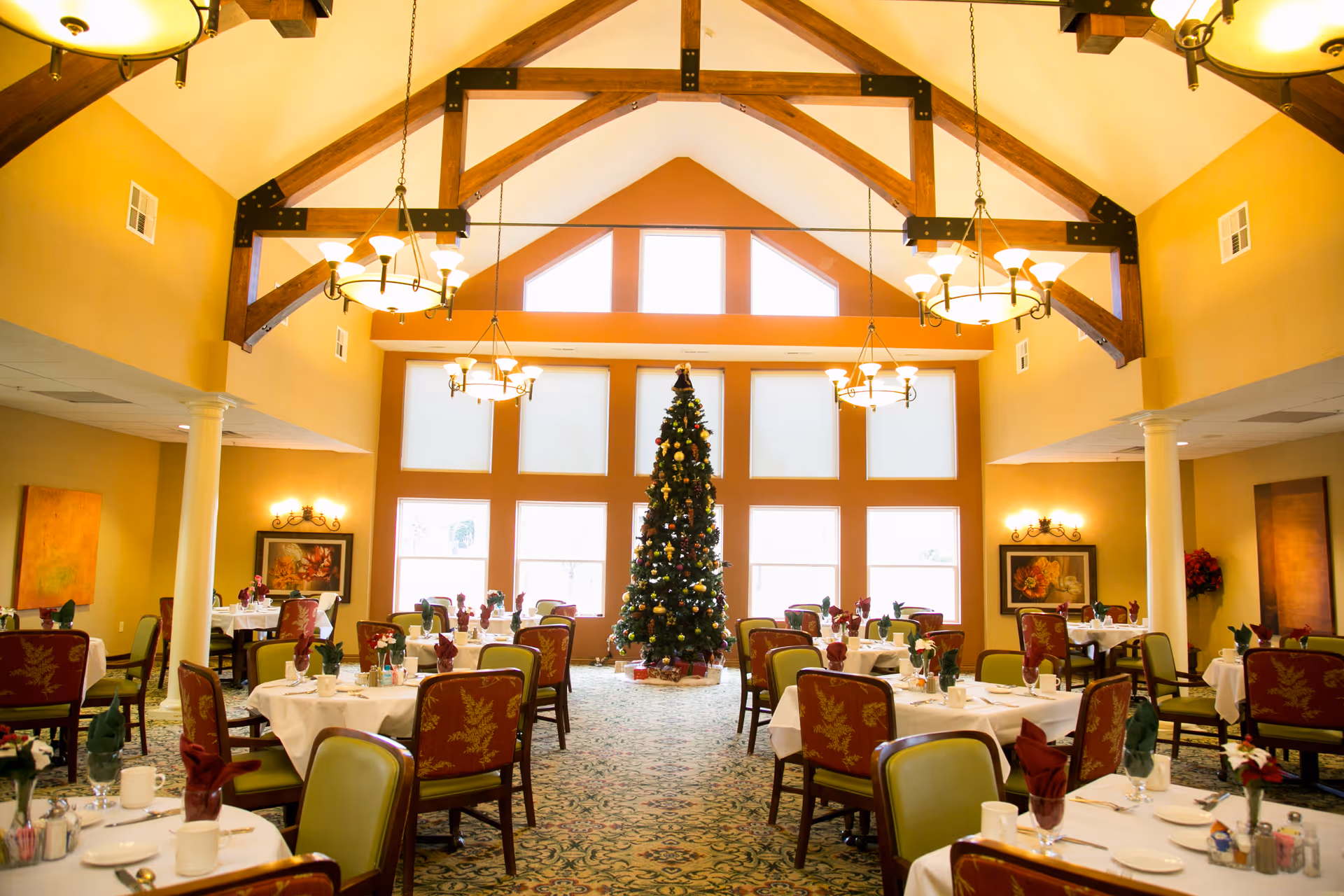 Spacious dining room with vaulted wooden beams, chandeliers, a decorated Christmas tree in the center, and multiple set dining tables.