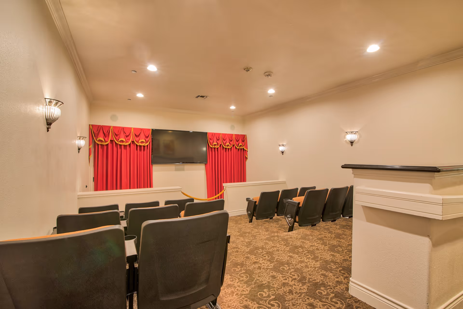 Small theater room with rows of black cushioned seats facing a large flat-screen TV mounted on a wall with red curtains. The room has beige walls, patterned carpet, and wall-mounted light fixtures.