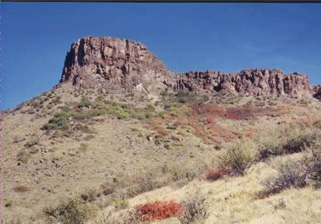 A rocky mesa with a flat top and steep sides rises above a dry, grassy landscape with scattered shrubs under a clear blue sky.