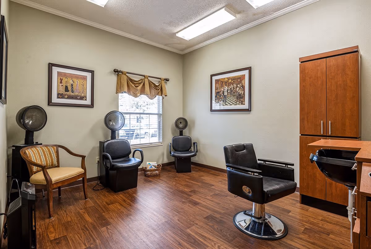 Interior of a hair salon room with wooden flooring, three black hair drying chairs with hooded dryers, a single salon styling chair, a wooden cabinet, a black wash basin, and two framed pictures on beige walls. A window with a brown valance allows natural light into the room.