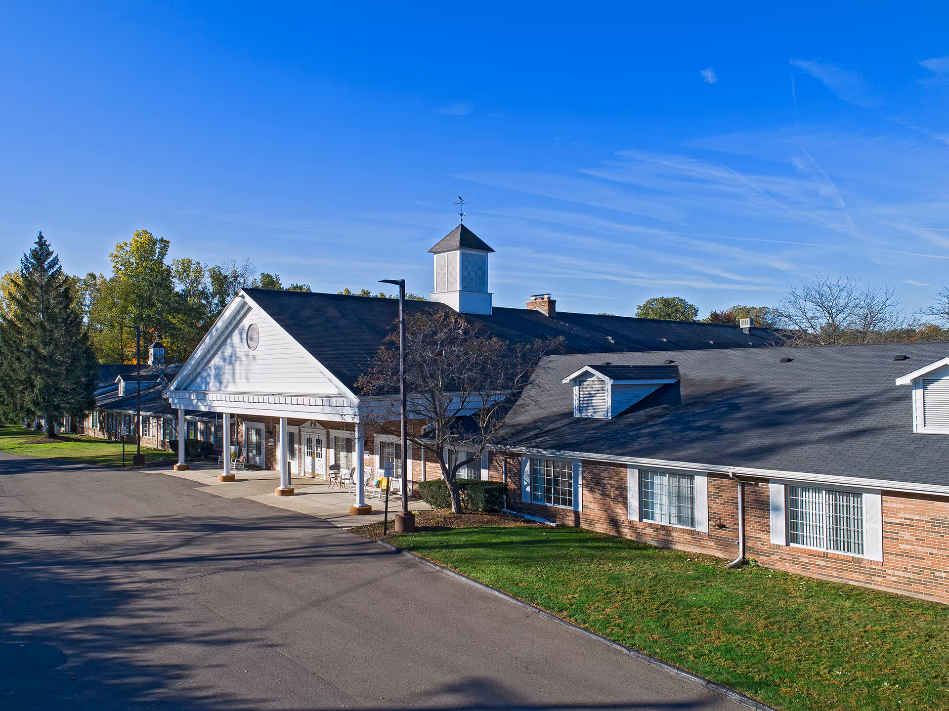Exterior view of a single-story brick building with white trim and a large covered entrance. The building has a dark shingled roof with dormer windows and a cupola on top. There is a paved driveway and green lawn with trees around the building under a clear blue sky.