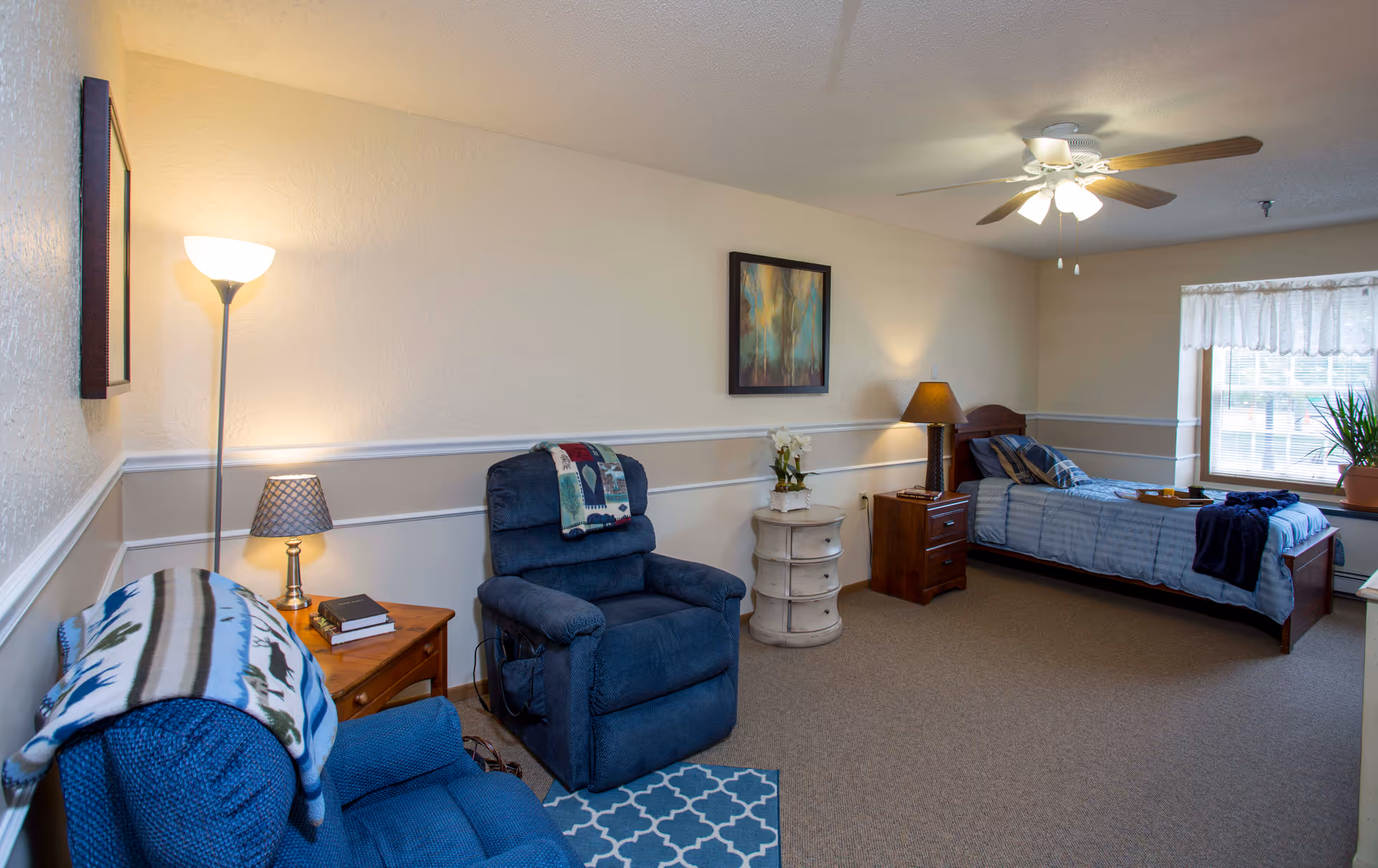 Well-lit senior living resident room with two blue recliners, side tables, and a single bed under a ceiling fan.