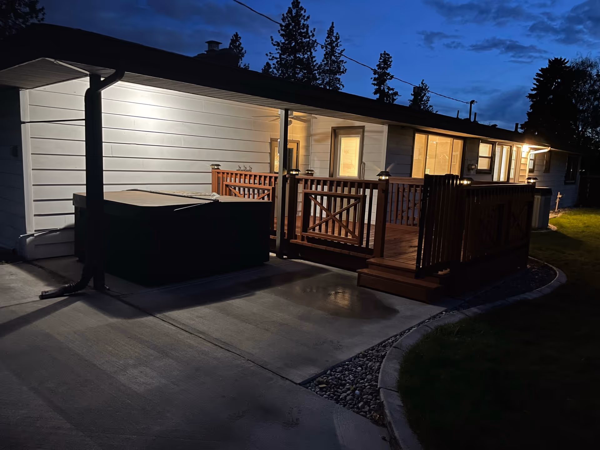 A lit wooden deck with railings and a covered hot tub attached to a single-story home's exterior at dusk.
