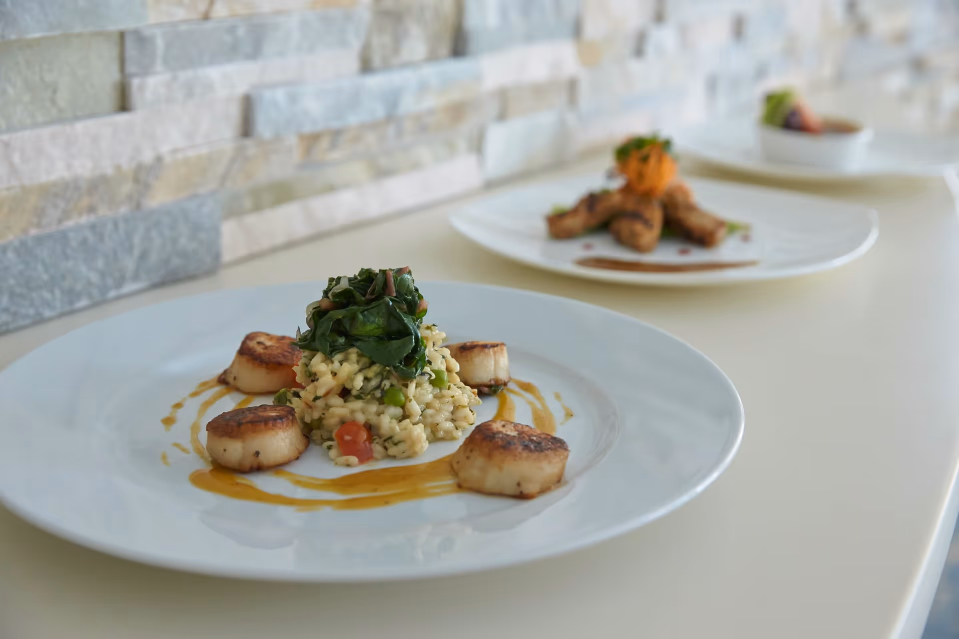 A close-up of a white plate with a gourmet dish featuring four seared scallops arranged around a mound of risotto topped with sautéed greens, with a drizzle of sauce on the plate. In the background, there are two more plates with different food items on a light-colored countertop against a stone wall.