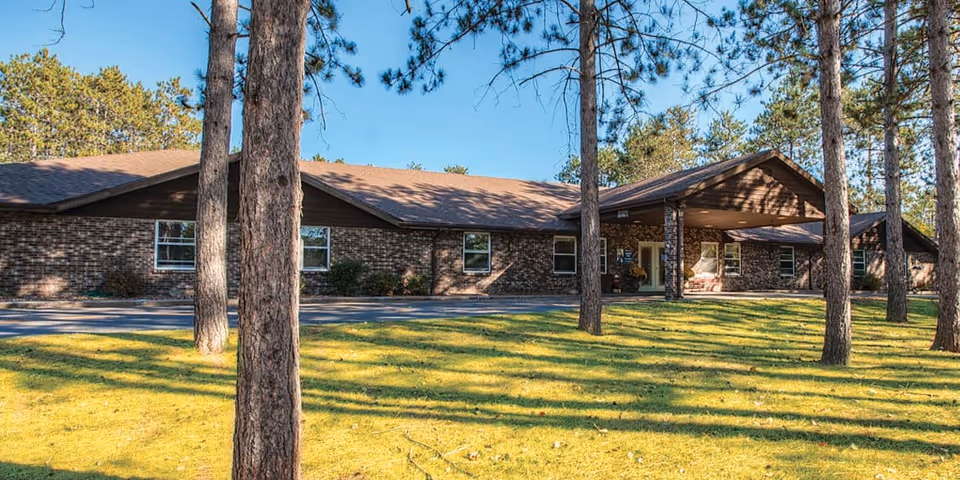 Single-story brick assisted living building with a covered entrance surrounded by pine trees and a grassy lawn.
