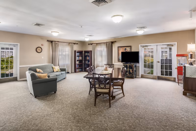 A spacious senior living common room with a seating area, dining table and chairs, TV, bookshelves, and double glass doors.