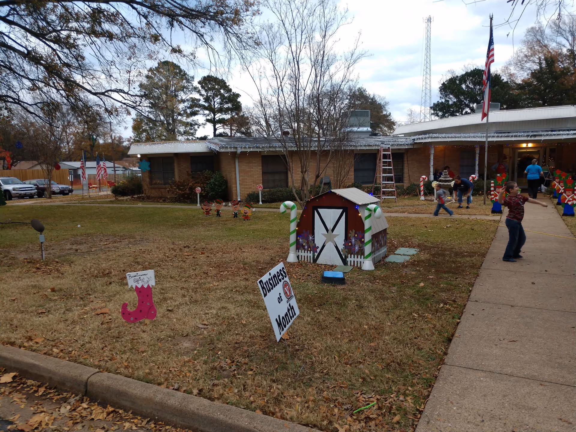 Front lawn of a single-story care facility decorated with holiday displays and a small play barn, with people near the entrance.