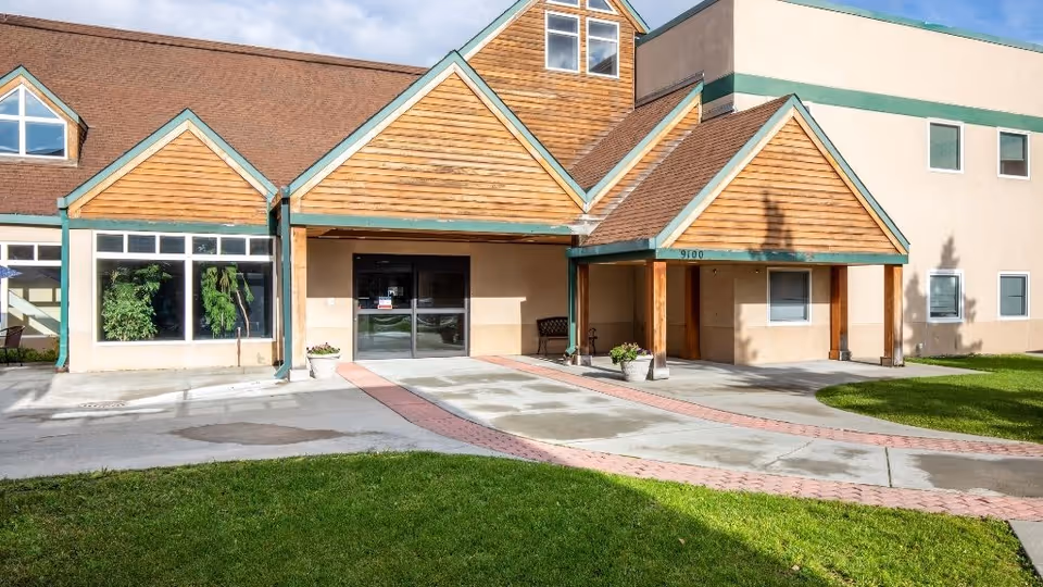 Front entrance of a senior living building with wooden gabled awnings, glass double doors, potted plants, and a brick-bordered driveway.