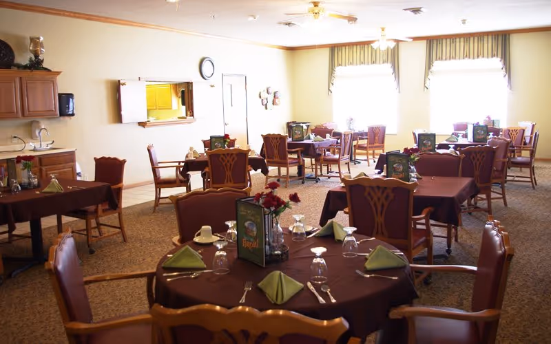 A dining room in a senior living facility with multiple tables covered in dark brown tablecloths, each set with green folded napkins, glasses, and silverware. The room has wooden chairs with cushioned seats, a carpeted floor, and large windows with valances letting in natural light. There is a small kitchenette area with cabinets and a sink on the left side of the room.