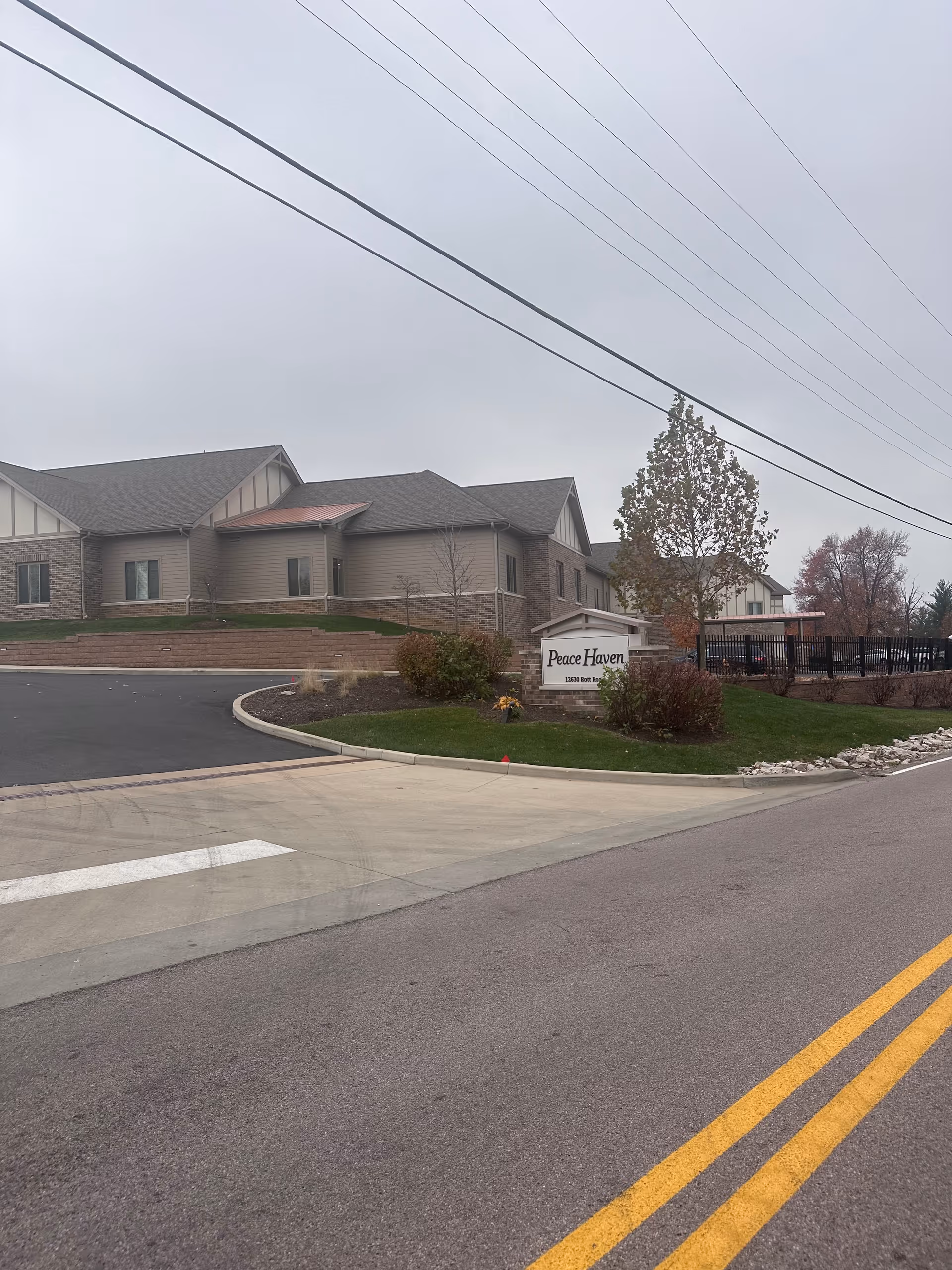 Front exterior of the Peace Haven senior living facility with its entrance sign, driveway, and surrounding landscaping under a cloudy sky.