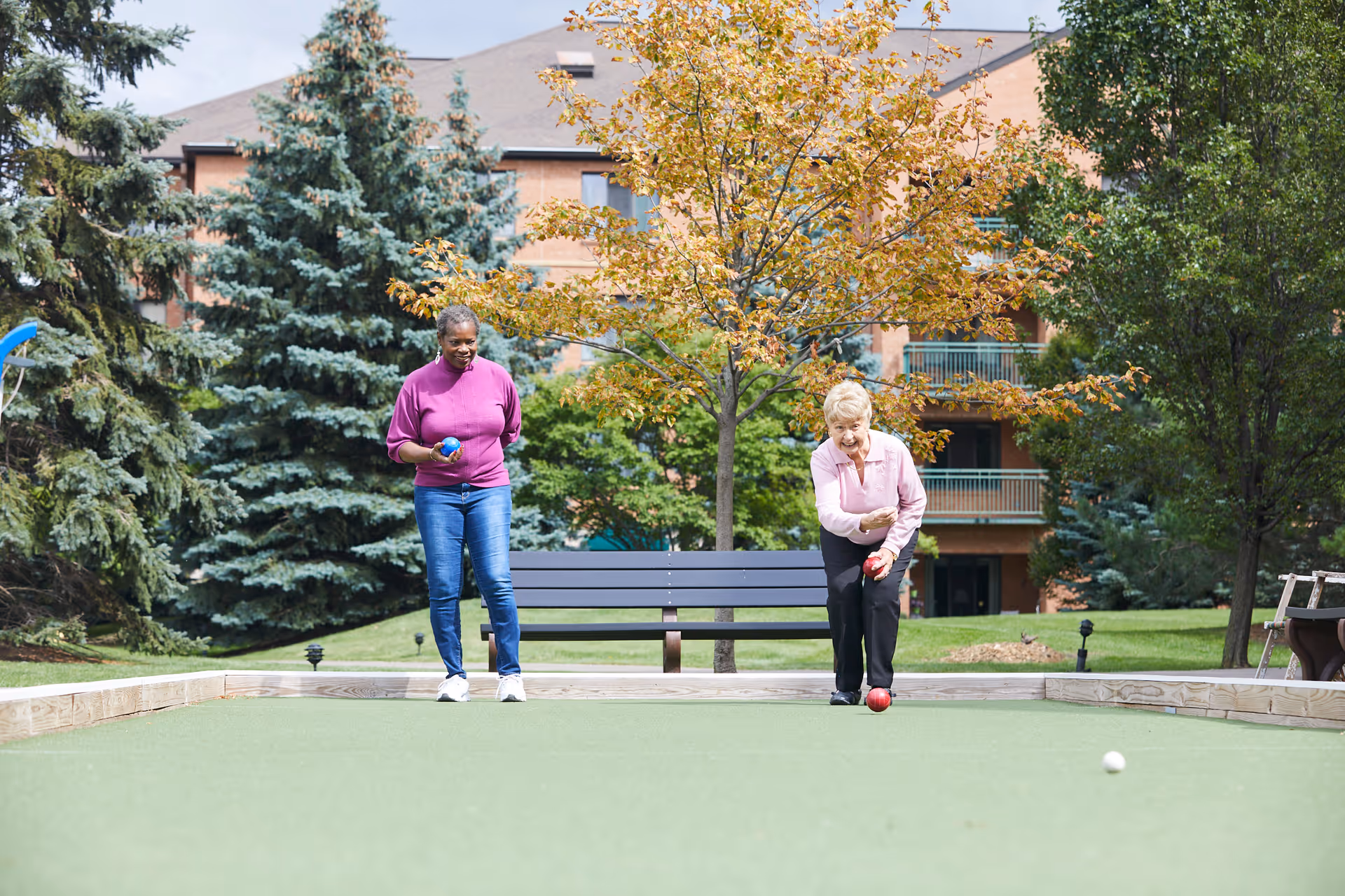 Two elderly women playing bocce ball on a green outdoor court surrounded by trees and a building in the background. One woman is in the process of rolling a red bocce ball while the other watches holding a blue ball.