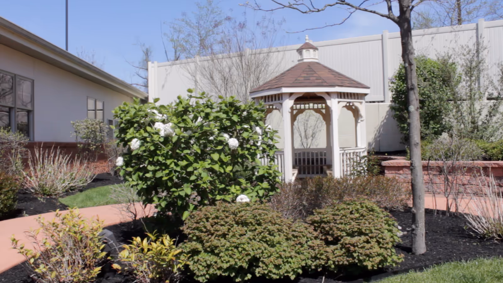 Outdoor garden area with a white gazebo surrounded by various bushes and plants, a tree, and a paved walkway. A building and a white fence are visible in the background under a clear blue sky.