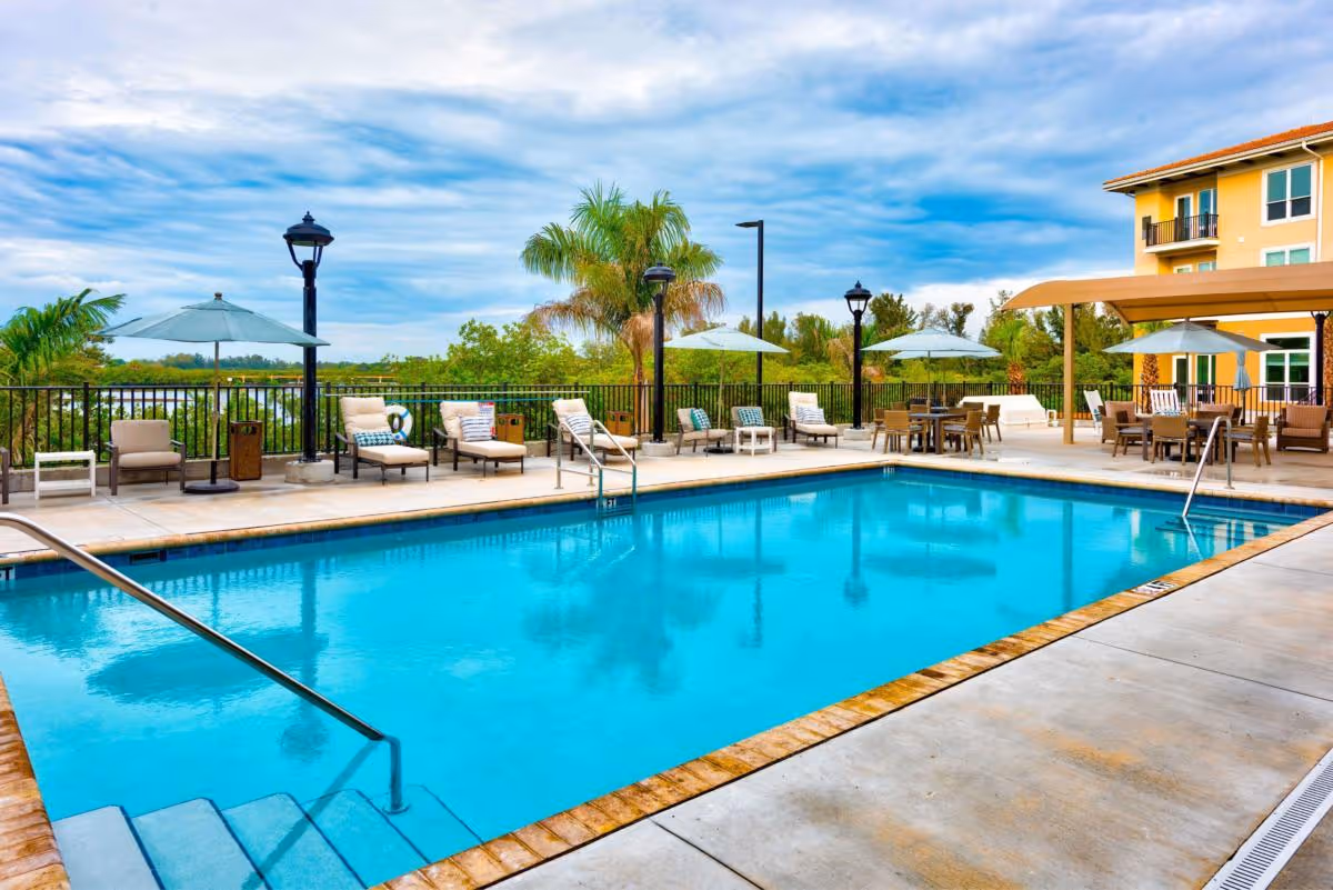 Outdoor swimming pool with lounge chairs, umbrellas, and a covered seating area beside a multi-story building.