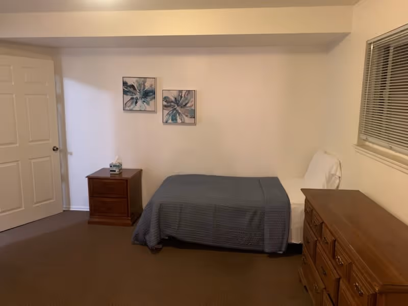 Small bedroom with a single bed covered by a blue quilt, wooden nightstand and dresser, two floral wall artworks, and a window with blinds.