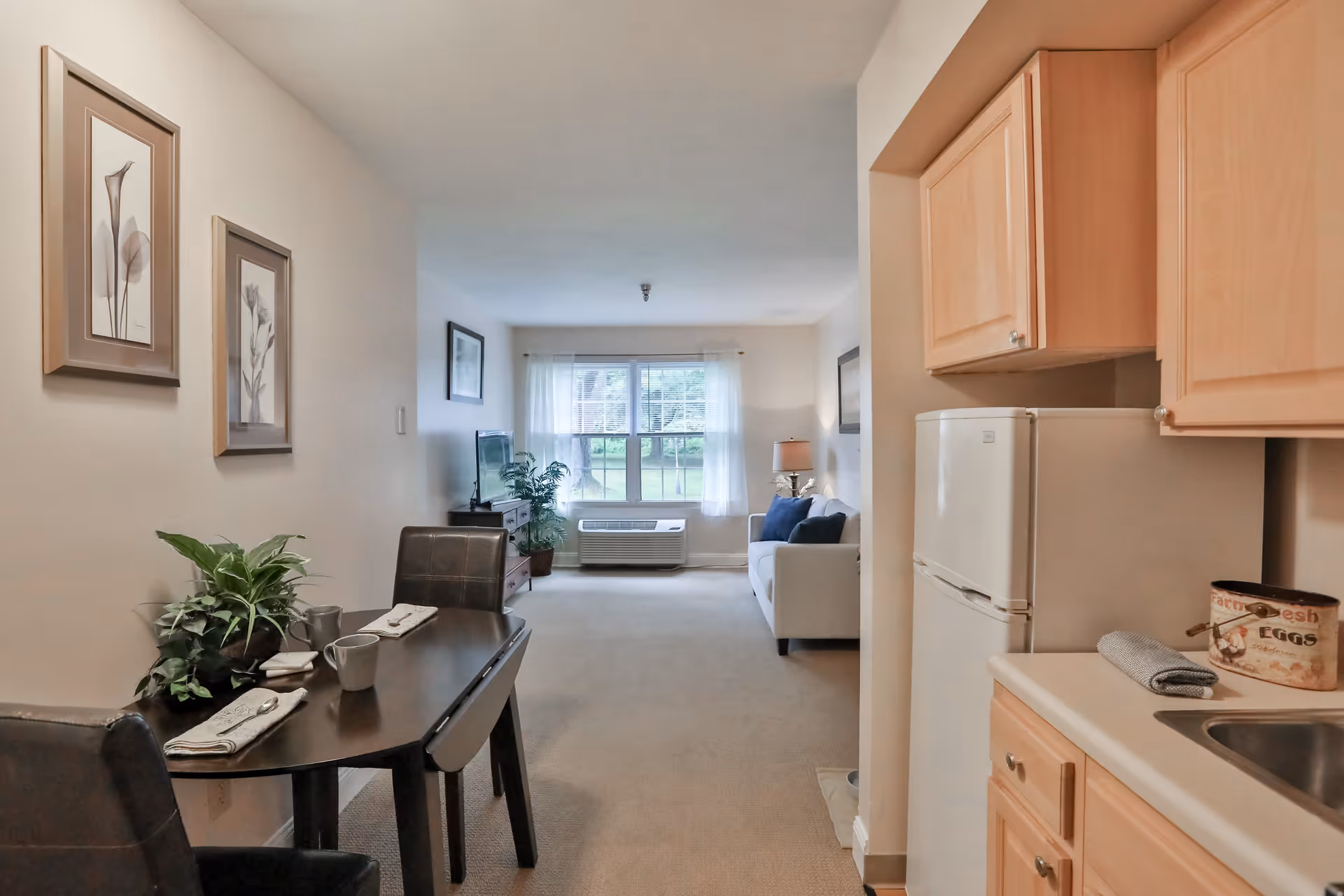 Interior view of a senior living apartment showing a small dining area with a table set for two, a kitchen with light wood cabinets and a white refrigerator, and a living room with a white sofa, blue cushions, a TV on a stand, and a large window with sheer curtains letting in natural light.