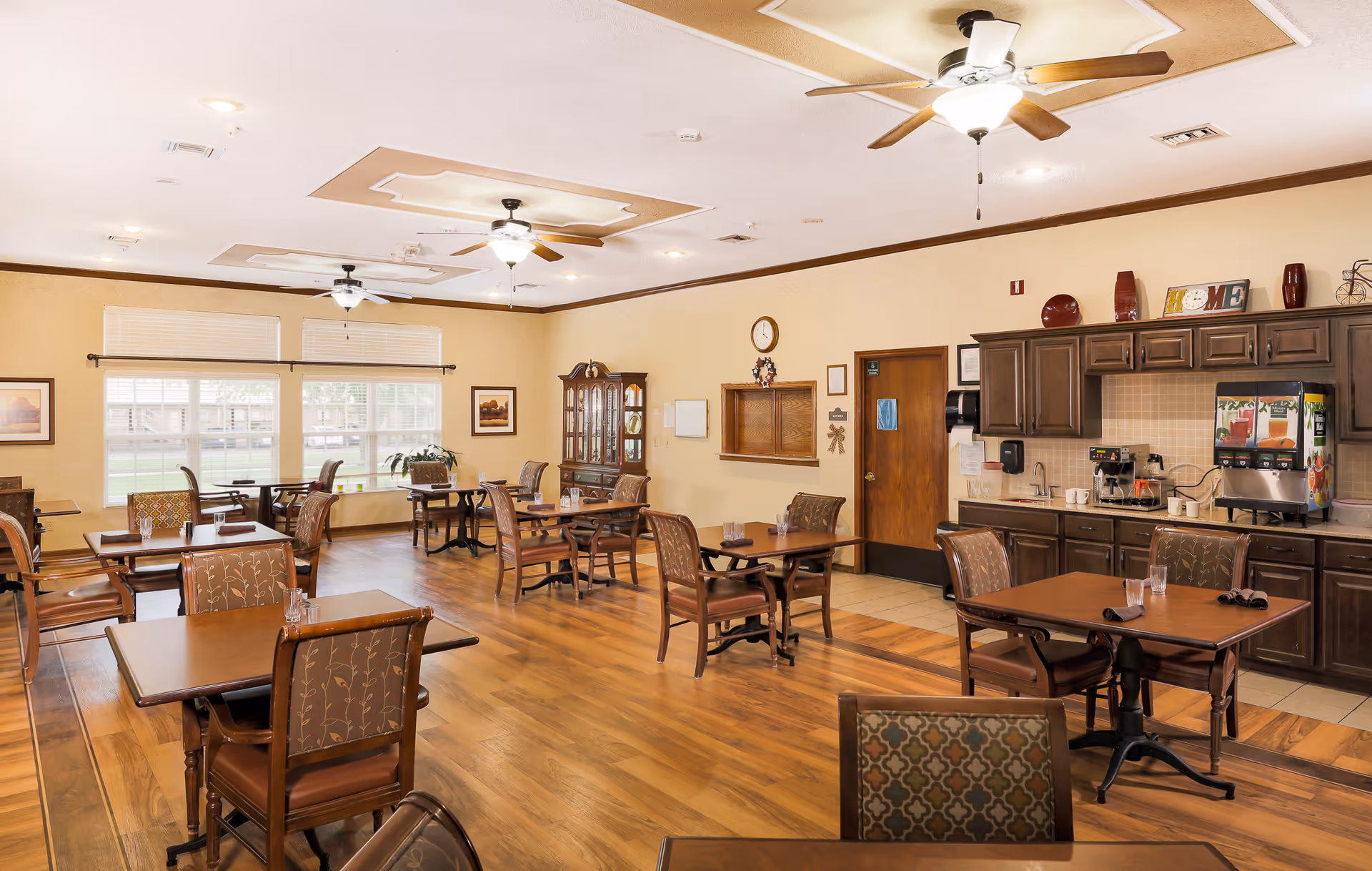 A spacious dining room with multiple wooden tables and chairs arranged neatly on a wooden floor. The room has large windows letting in natural light, ceiling fans with lights, and a counter area with beverage dispensers and cabinets. The walls are decorated with framed pictures and a clock.