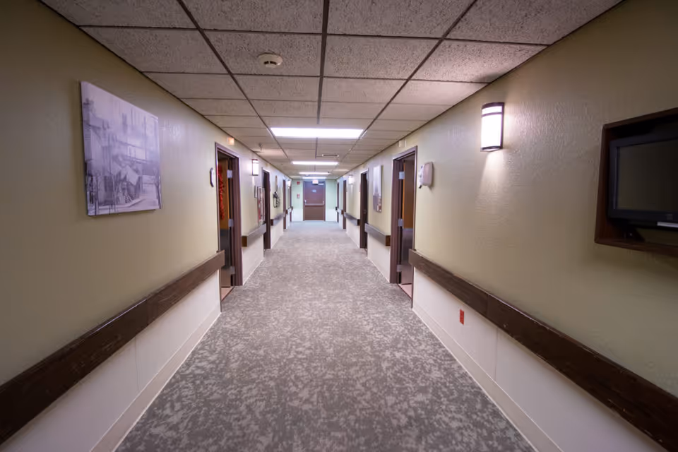 A long, well-lit hallway in a healthcare facility with beige walls, carpeted floor, and wooden handrails on both sides. Several open doorways lead to rooms along the corridor, and there are wall-mounted lights and framed pictures on the walls.