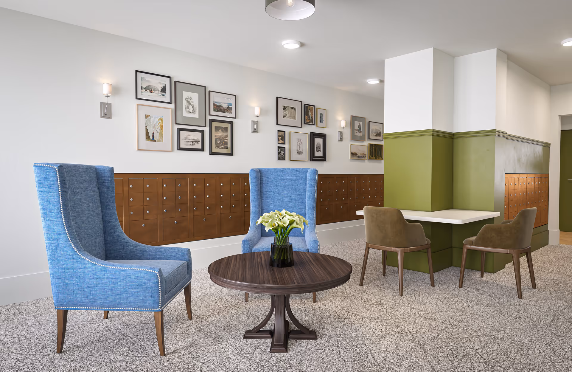 A cozy seating area in a senior living facility featuring two blue upholstered armchairs around a round wooden table with a vase of white flowers. Behind the chairs is a wall with multiple framed artworks and a row of brown mailboxes. To the right, there is a small table with two brown chairs against a green and white half-wall.