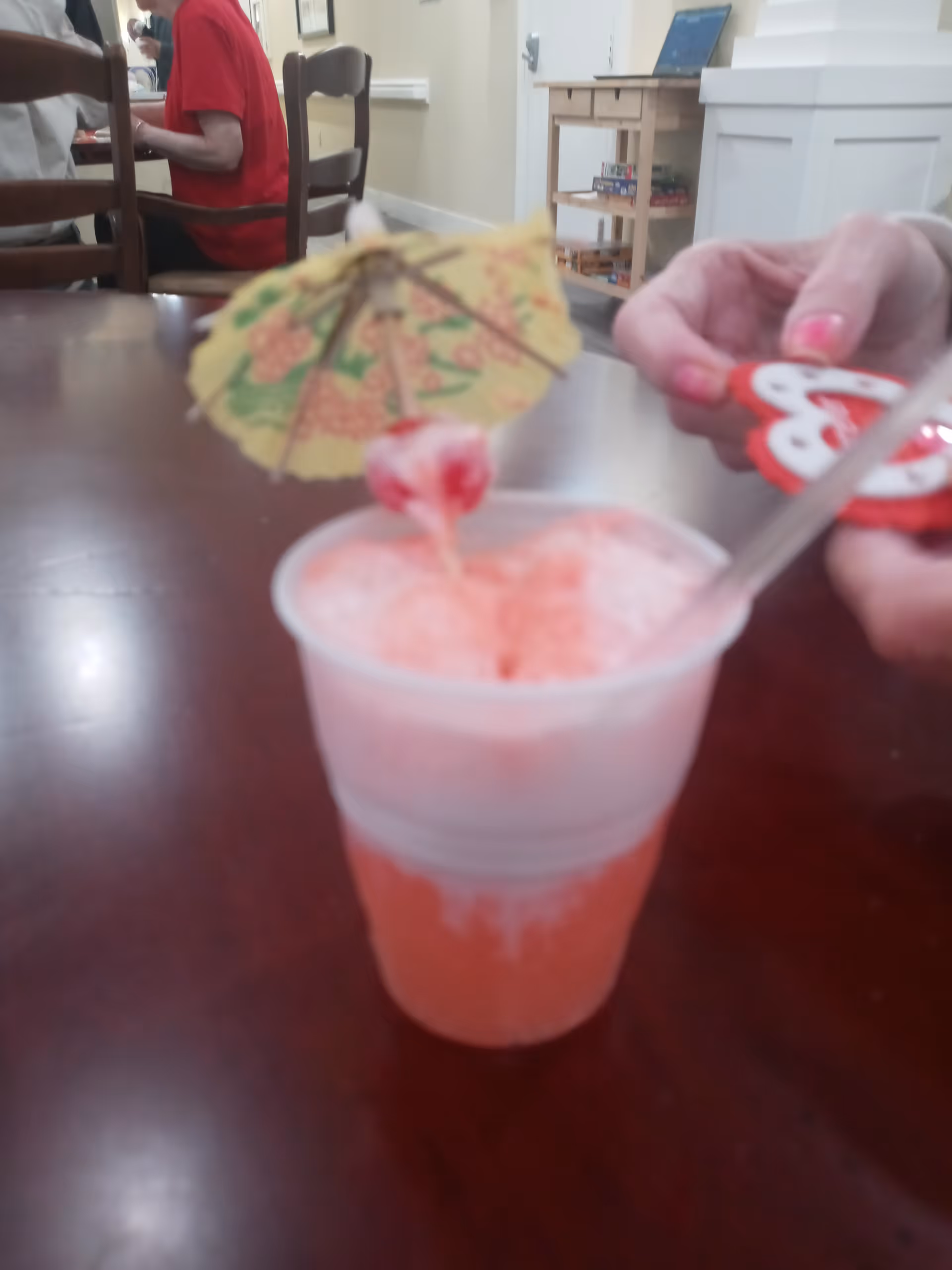 A close-up of a pink frozen drink in a clear plastic cup with a decorative paper umbrella and a cherry on top, placed on a wooden table. In the background, a person wearing a red shirt is seated at the table, and another person's hands with pink nail polish are holding a red and white object. The setting appears to be an indoor common area with chairs and a small shelf with board games.
