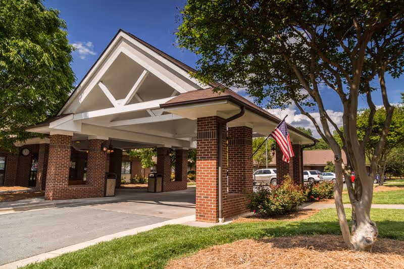 Entrance area of Oak Forest Health and Rehabilitation Center featuring a covered drop-off with brick pillars, an American flag, trees, and parked cars under a partly cloudy sky.