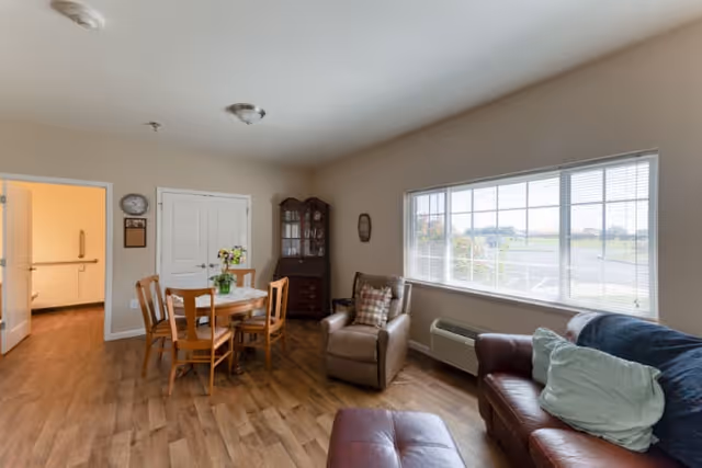 A bright and cozy living area in Cedarhurst of Breese featuring a wooden dining table with four chairs, a cushioned armchair with a plaid pillow, a leather sofa with pillows, a wooden cabinet with glass doors, and a large window letting in natural light overlooking an outdoor parking area.