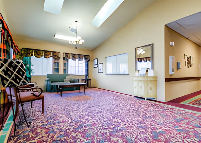 Interior view of a living room area with a patterned carpet, a green sofa, a wooden coffee table, chairs, and windows with valances. The room has beige walls, a skylight, and a hallway visible to the right.