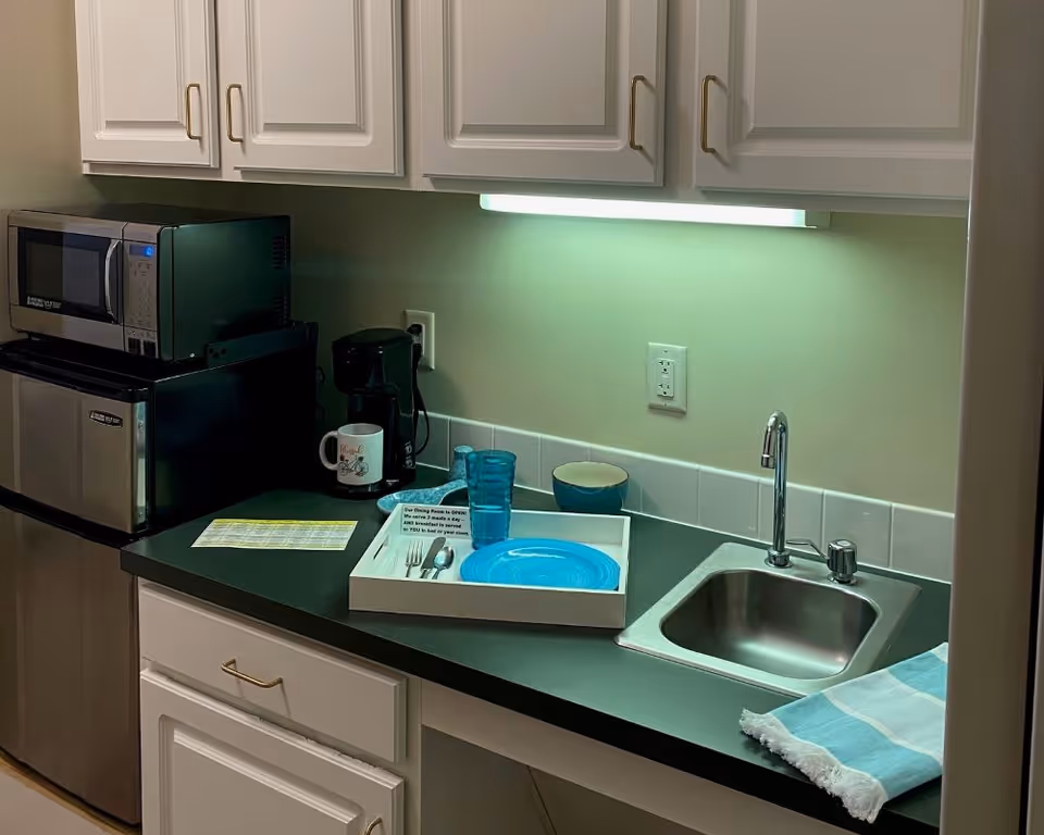 A small kitchenette area with white cabinets, a black countertop, a stainless steel sink with a faucet, a microwave, a mini refrigerator, a coffee maker with a mug, a tray with blue plates and cups, utensils, and a folded blue and white striped towel.