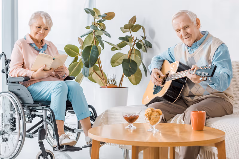 An elderly woman in a wheelchair reading a book and an elderly man sitting on a couch playing an acoustic guitar in a bright living room with a large potted plant and a wooden coffee table with snacks and a mug.