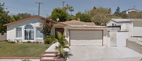 Single-story residential building with a white exterior, a two-car garage, and a front yard with green grass and a small palm tree. There are bushes and flowers near the front window, and a concrete driveway leading to the garage. The sky is clear and blue.