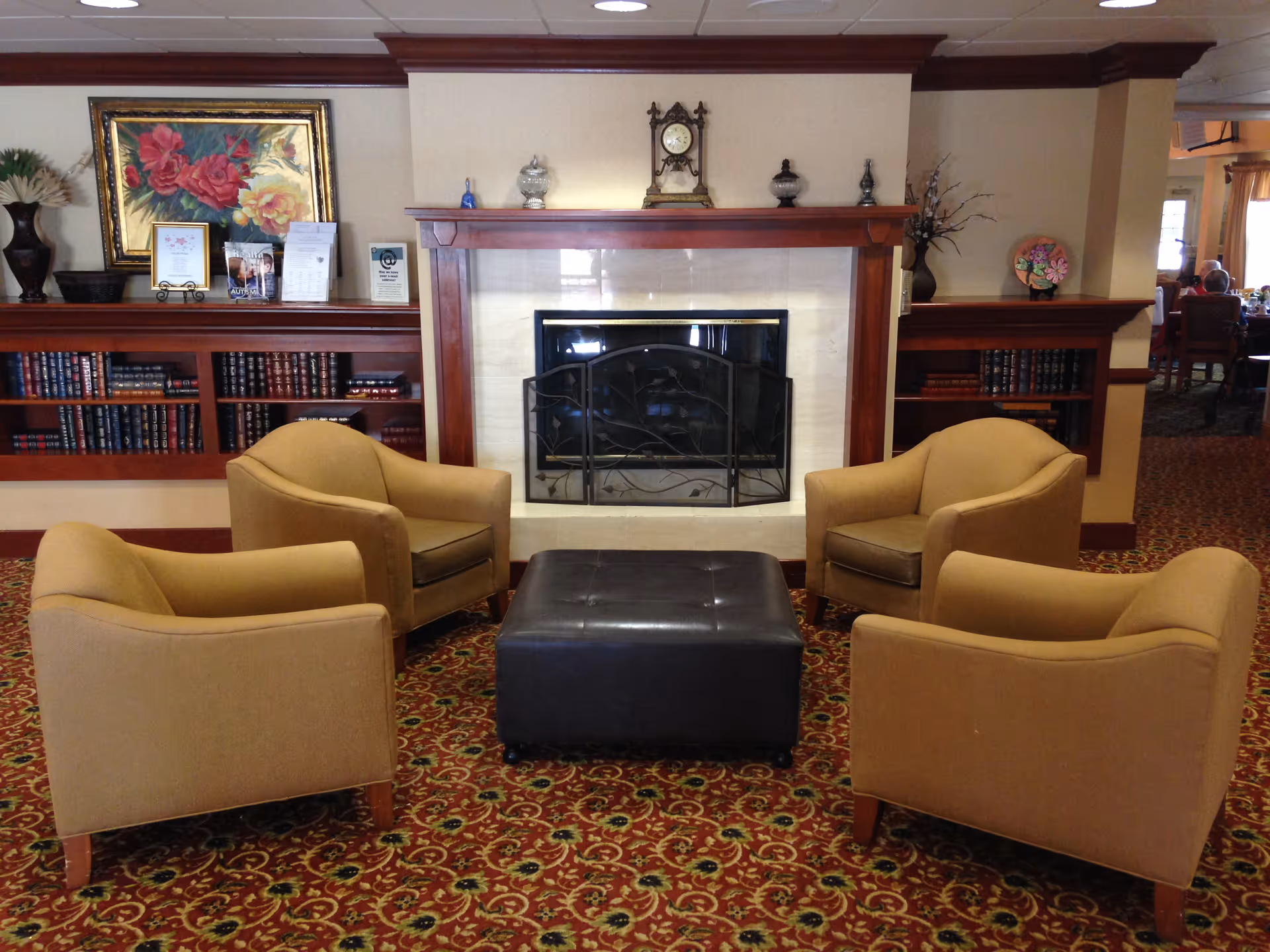 A cozy seating area with four beige armchairs arranged around a black ottoman in front of a fireplace with a wooden mantel. The mantel holds decorative items including a clock and small vases. Behind the seating area are built-in wooden bookshelves filled with books and decorative pieces, and a framed floral painting is displayed on the wall. The room has a patterned carpet and warm lighting.