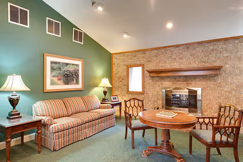 A cozy living room with a striped sofa, two wooden side tables each with a lamp, a round wooden table with a chessboard on it, and three wooden chairs. The room has a green carpet, a green accent wall with three vents and a framed picture, and a brick wall with a fireplace and a small window.